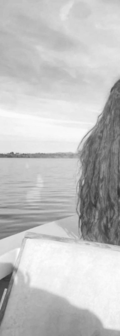 A black and white photo shows a woman sitting on a boat, seen from behind. Her very long hair flows down her back.